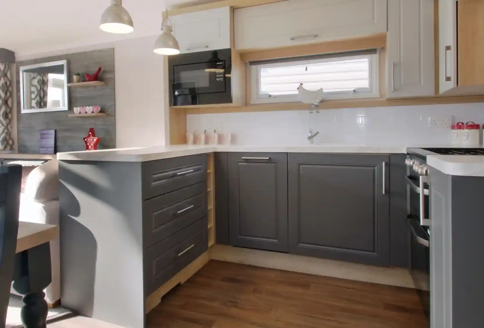 A modern kitchen featuring gray and white cabinetry, a countertop with a glossy finish, and silver pendant lights. A microwave is mounted above the counter, and wooden flooring extends throughout the space. Shelves in the background display decorative items.