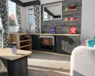 A cozy living room featuring a textured wall with wooden shelves displaying decorative items. A modern electric fireplace sits in a sleek dark cabinet, and a large mirror reflects natural light. Soft curtains frame the windows, while a light-colored couch and a coffee table complete the warm atmosphere.