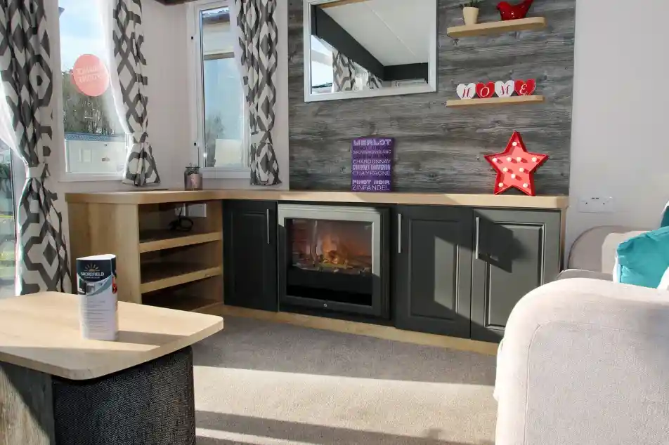 A cozy living room featuring a textured wall with wooden shelves displaying decorative items. A modern electric fireplace sits in a sleek dark cabinet, and a large mirror reflects natural light. Soft curtains frame the windows, while a light-colored couch and a coffee table complete the warm atmosphere.