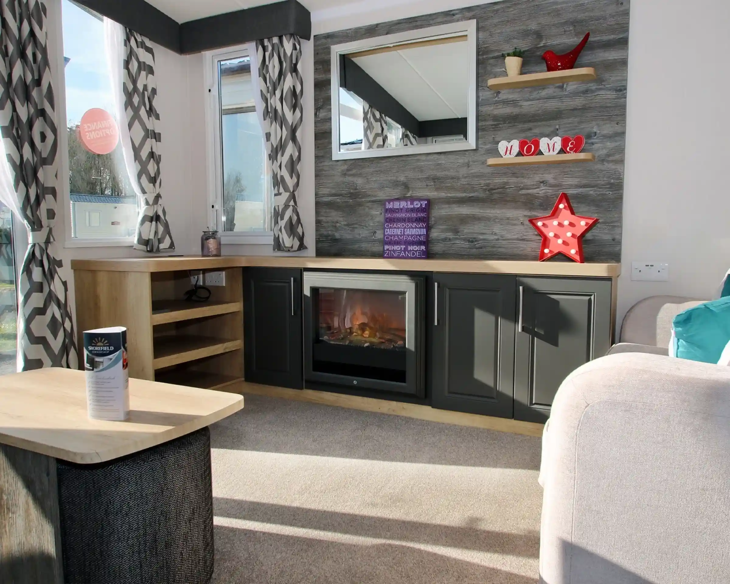 A cozy living room featuring a textured wall with wooden shelves displaying decorative items. A modern electric fireplace sits in a sleek dark cabinet, and a large mirror reflects natural light. Soft curtains frame the windows, while a light-colored couch and a coffee table complete the warm atmosphere.