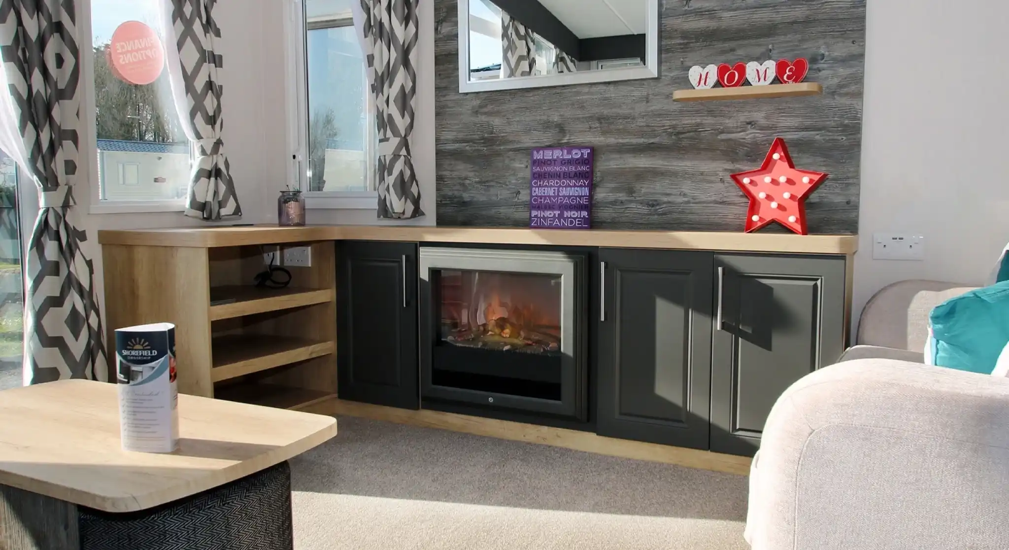 A cozy living room featuring a textured wall with wooden shelves displaying decorative items. A modern electric fireplace sits in a sleek dark cabinet, and a large mirror reflects natural light. Soft curtains frame the windows, while a light-colored couch and a coffee table complete the warm atmosphere.