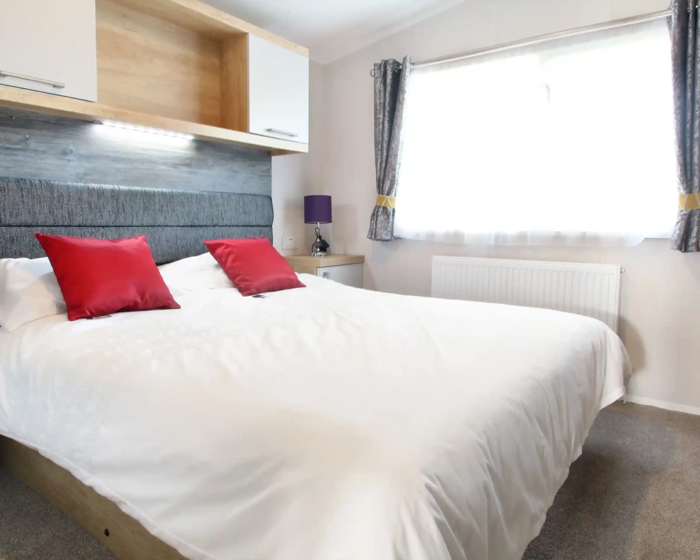 A well-lit bedroom featuring a large white bed with red decorative pillows. The headboard is gray, and there is a wooden shelf above the bed. A purple lamp sits on a nightstand beside the bed, and a window with sheer curtains lets in natural light.
