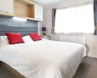A well-lit bedroom featuring a large white bed with red decorative pillows. The headboard is gray, and there is a wooden shelf above the bed. A purple lamp sits on a nightstand beside the bed, and a window with sheer curtains lets in natural light.