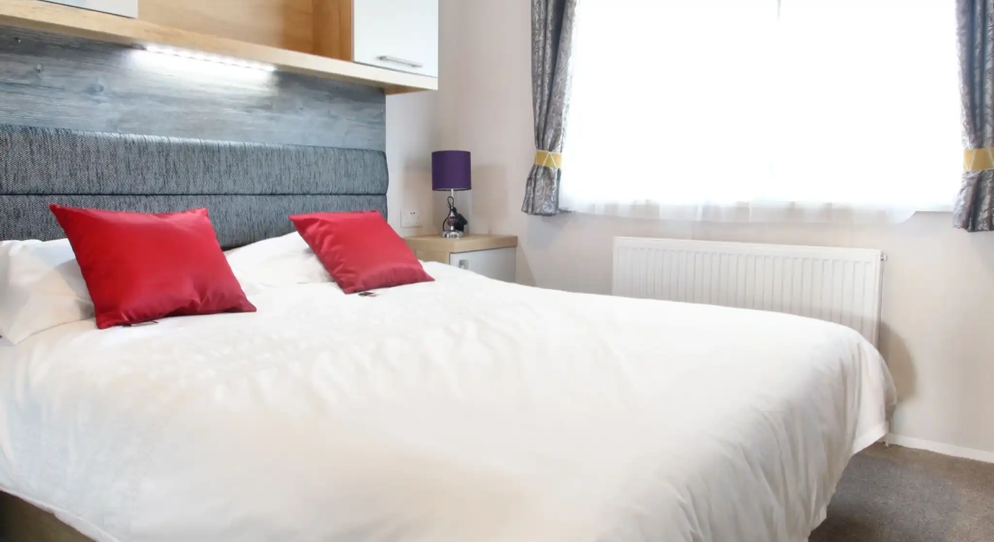 A well-lit bedroom featuring a large white bed with red decorative pillows. The headboard is gray, and there is a wooden shelf above the bed. A purple lamp sits on a nightstand beside the bed, and a window with sheer curtains lets in natural light.