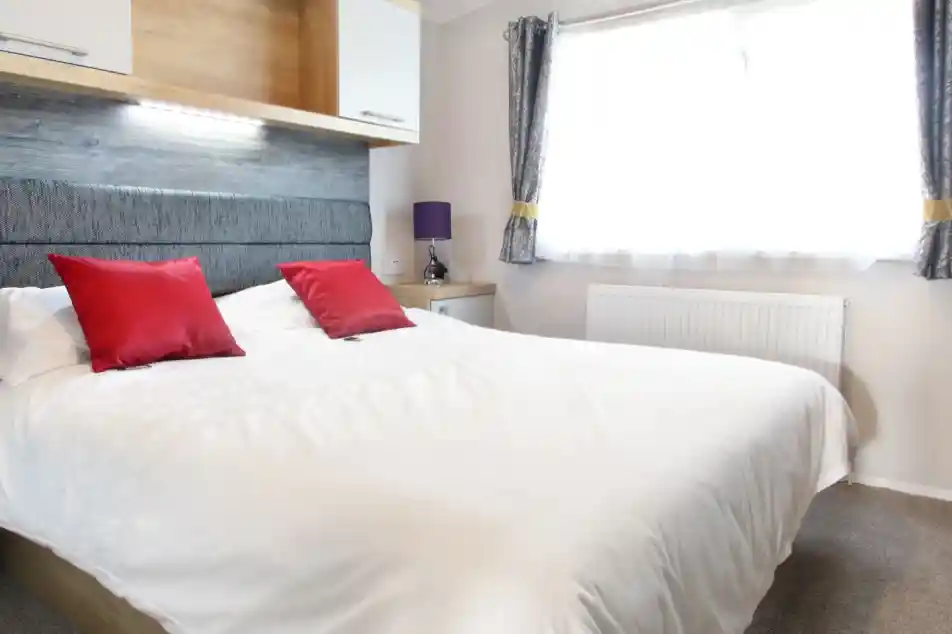 A well-lit bedroom featuring a large white bed with red decorative pillows. The headboard is gray, and there is a wooden shelf above the bed. A purple lamp sits on a nightstand beside the bed, and a window with sheer curtains lets in natural light.