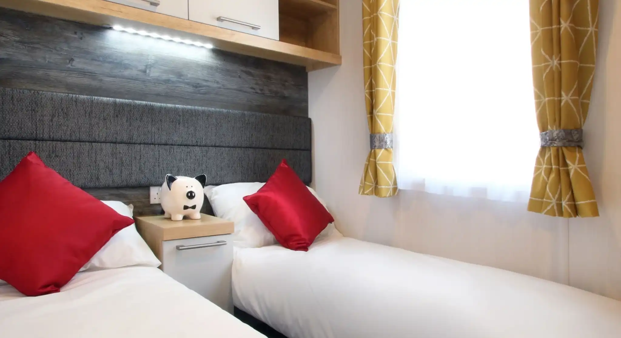 A cozy bedroom featuring two single beds with white linens and red accent pillows. A small nightstand with a stuffed panda is between the beds. Bright yellow curtains frame a window, allowing natural light to enter.