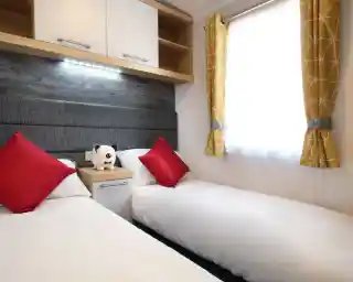 A cozy bedroom featuring two single beds with white linens and red accent pillows. A small nightstand with a stuffed panda is between the beds. Bright yellow curtains frame a window, allowing natural light to enter.