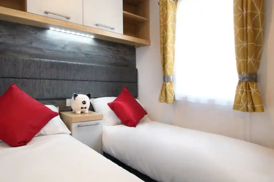 A cozy bedroom featuring two single beds with white linens and red accent pillows. A small nightstand with a stuffed panda is between the beds. Bright yellow curtains frame a window, allowing natural light to enter.