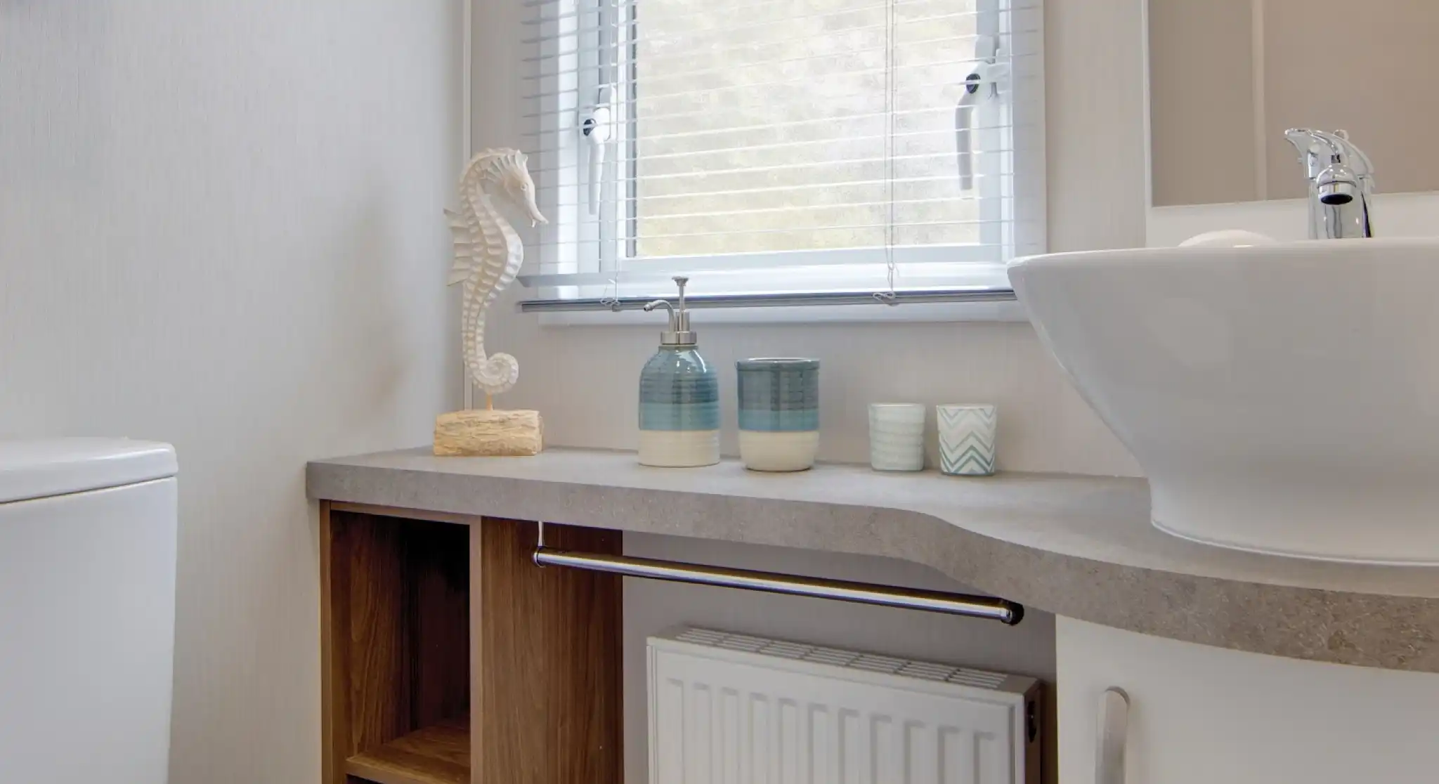 Modern bathroom with a white sink, soap dispensers, decorative seahorse, and towels on shelves below a window with blinds.