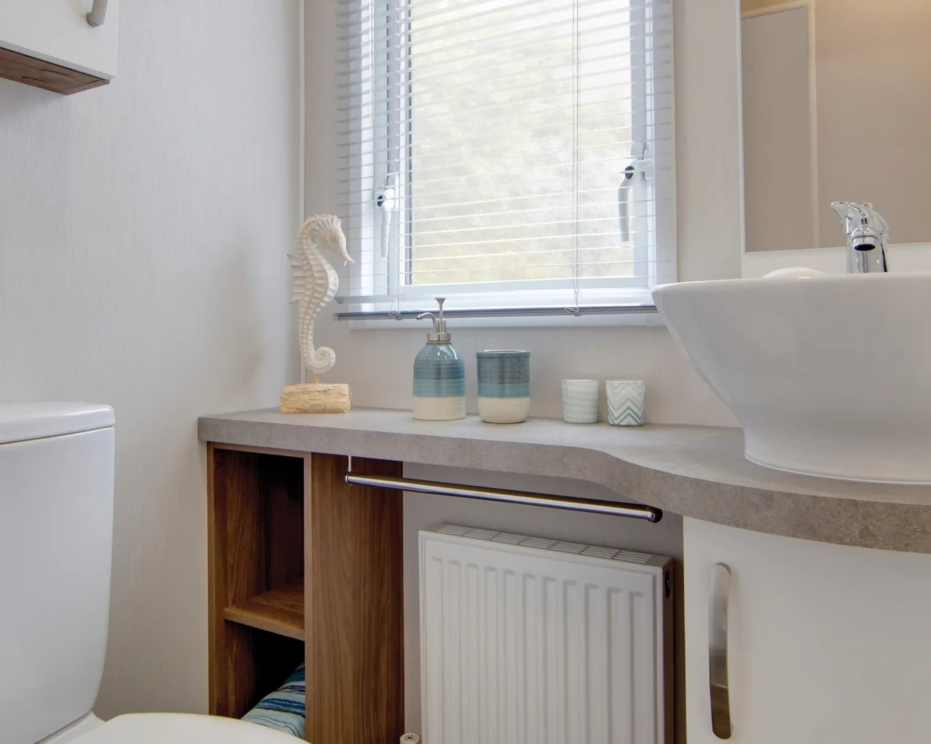 Modern bathroom with a white sink, soap dispensers, decorative seahorse, and towels on shelves below a window with blinds.