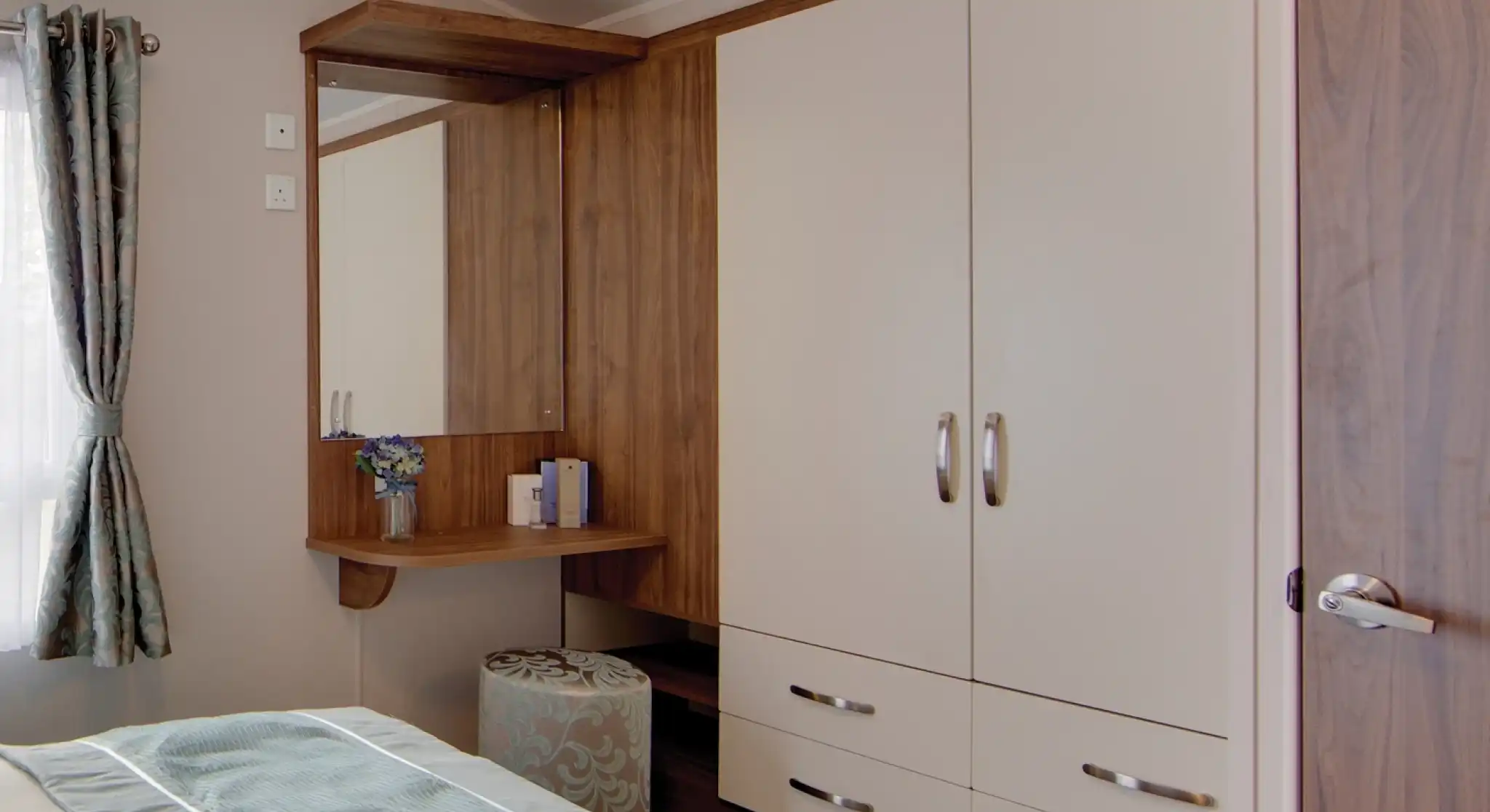 A bedroom corner with built-in cream wardrobes featuring wood trim and silver handles. Next to the wardrobe is a wooden vanity with a mirror, a small vase of flowers, and some items neatly placed on the surface. A round upholstered stool with a blue and beige pattern sits beneath the vanity. A window with patterned curtains allows natural light into the room, and part of the bed with light blue bedding is visible in the foreground.