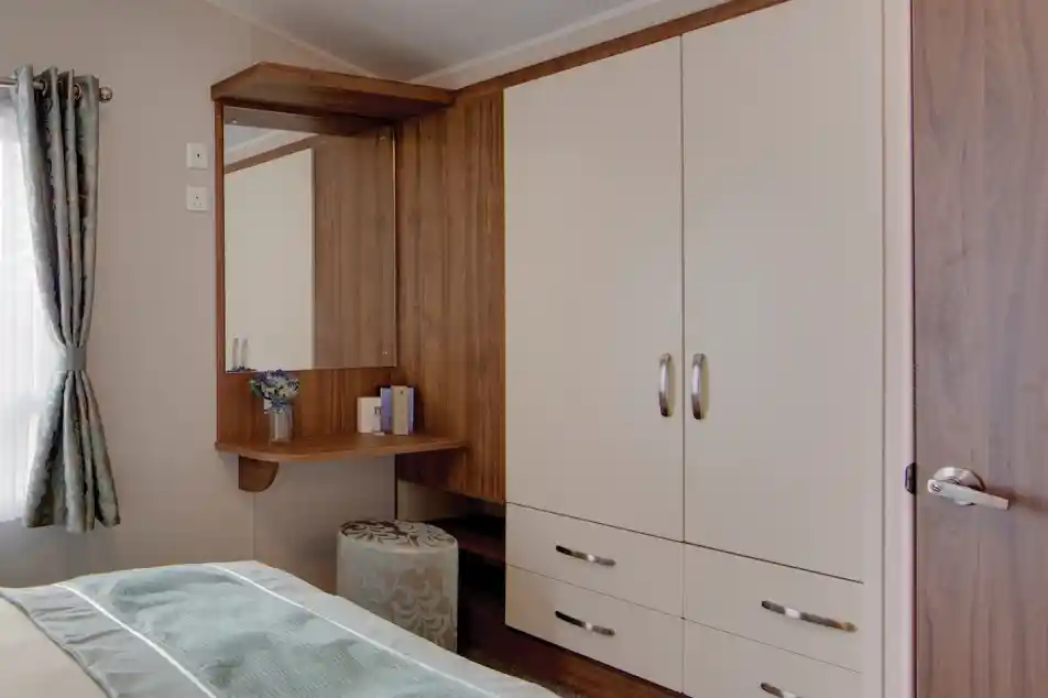 A bedroom corner with built-in cream wardrobes featuring wood trim and silver handles. Next to the wardrobe is a wooden vanity with a mirror, a small vase of flowers, and some items neatly placed on the surface. A round upholstered stool with a blue and beige pattern sits beneath the vanity. A window with patterned curtains allows natural light into the room, and part of the bed with light blue bedding is visible in the foreground.
