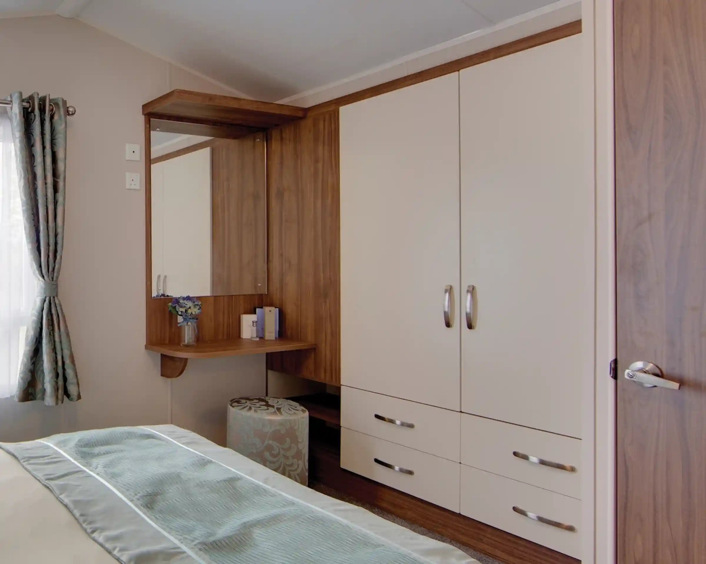 A bedroom corner with built-in cream wardrobes featuring wood trim and silver handles. Next to the wardrobe is a wooden vanity with a mirror, a small vase of flowers, and some items neatly placed on the surface. A round upholstered stool with a blue and beige pattern sits beneath the vanity. A window with patterned curtains allows natural light into the room, and part of the bed with light blue bedding is visible in the foreground.