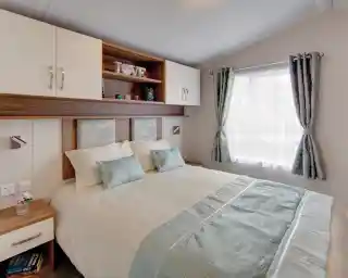 A neatly arranged bedroom with a double bed covered in cream and light blue bedding. The bed has two pillows and two decorative cushions, with a light blue patterned headboard. Above the bed are wall-mounted cabinets and open shelves holding decor items, including candles and a decorative piece spelling “LOVE.” To the right, a window with sheer white curtains and light blue patterned drapes lets in natural light. A wooden bedside table with a drawer and books is positioned to the left of the bed.