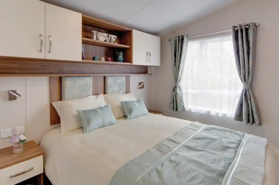 A neatly arranged bedroom with a double bed covered in cream and light blue bedding. The bed has two pillows and two decorative cushions, with a light blue patterned headboard. Above the bed are wall-mounted cabinets and open shelves holding decor items, including candles and a decorative piece spelling “LOVE.” To the right, a window with sheer white curtains and light blue patterned drapes lets in natural light. A wooden bedside table with a drawer and books is positioned to the left of the bed.
