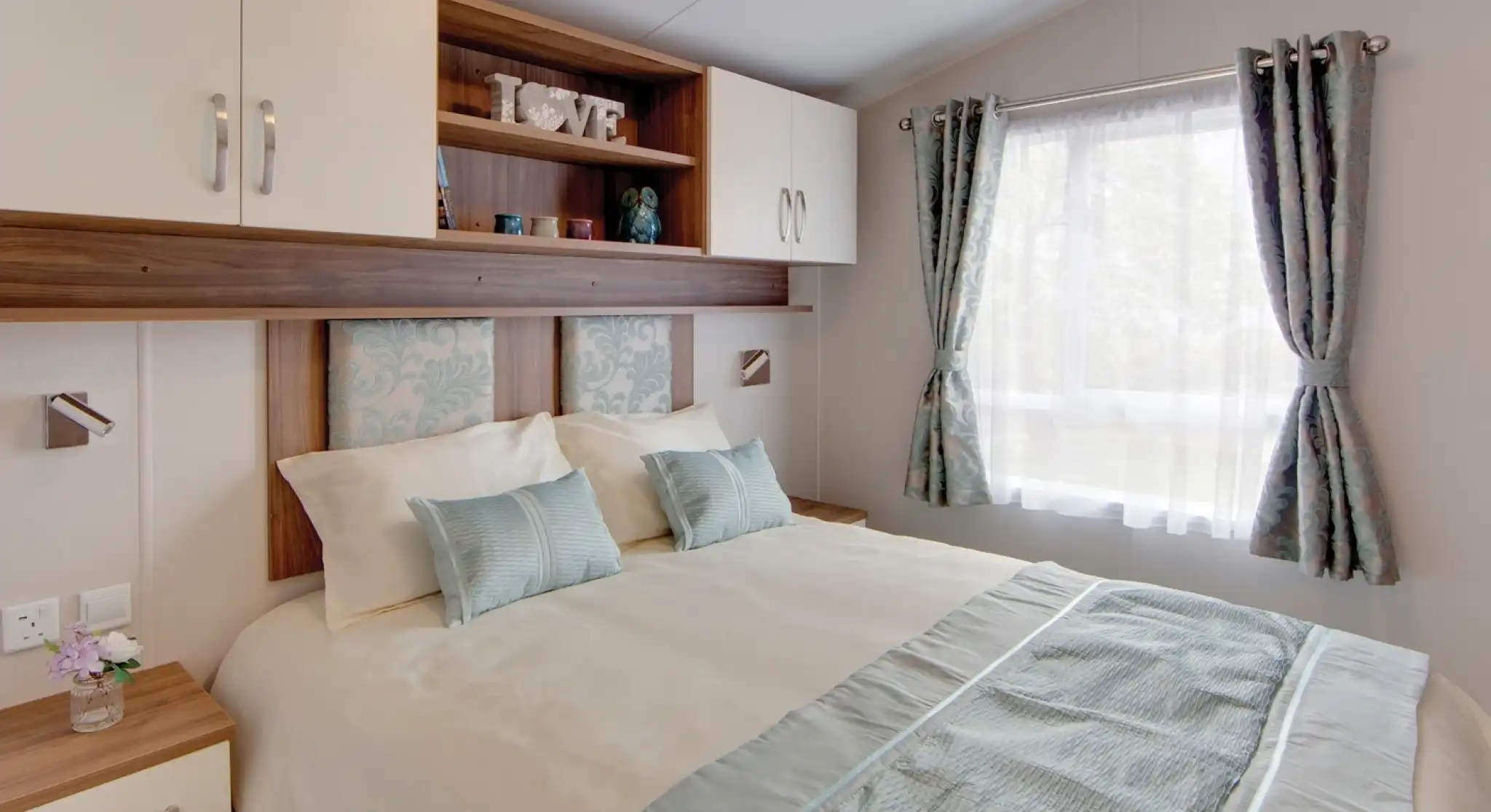 A neatly arranged bedroom with a double bed covered in cream and light blue bedding. The bed has two pillows and two decorative cushions, with a light blue patterned headboard. Above the bed are wall-mounted cabinets and open shelves holding decor items, including candles and a decorative piece spelling “LOVE.” To the right, a window with sheer white curtains and light blue patterned drapes lets in natural light. A wooden bedside table with a drawer and books is positioned to the left of the bed.