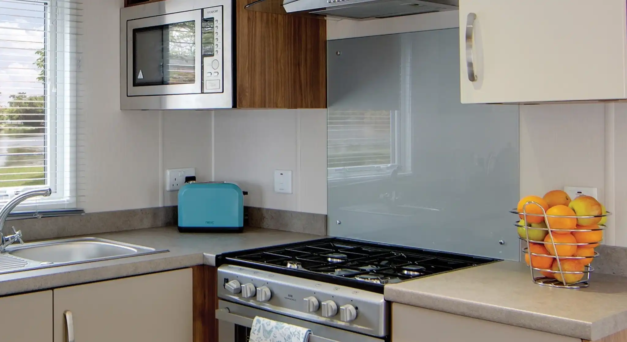 A modern kitchen counter with a stainless steel gas stove, a blue toaster, a microwave, and a basket of oranges.
