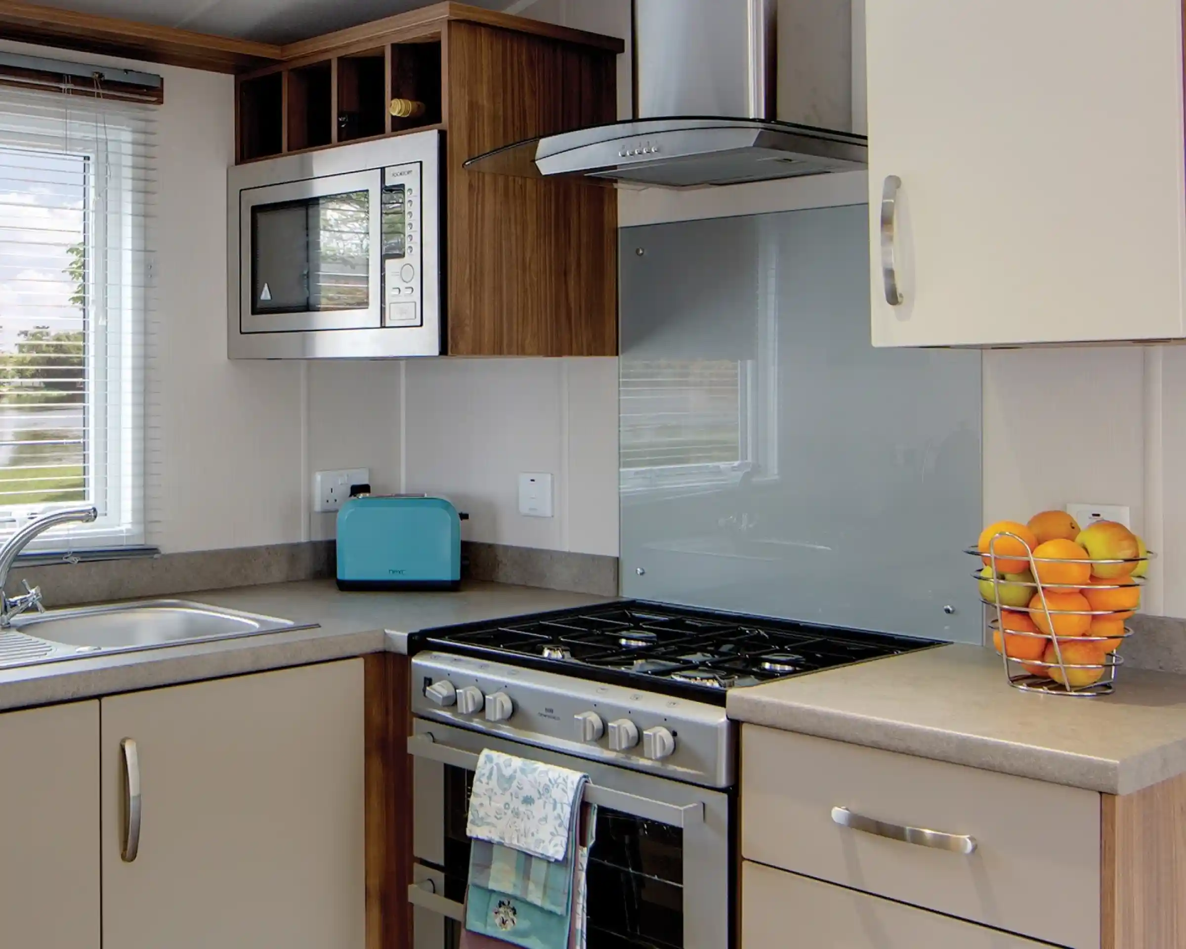 A modern kitchen counter with a stainless steel gas stove, a blue toaster, a microwave, and a basket of oranges.