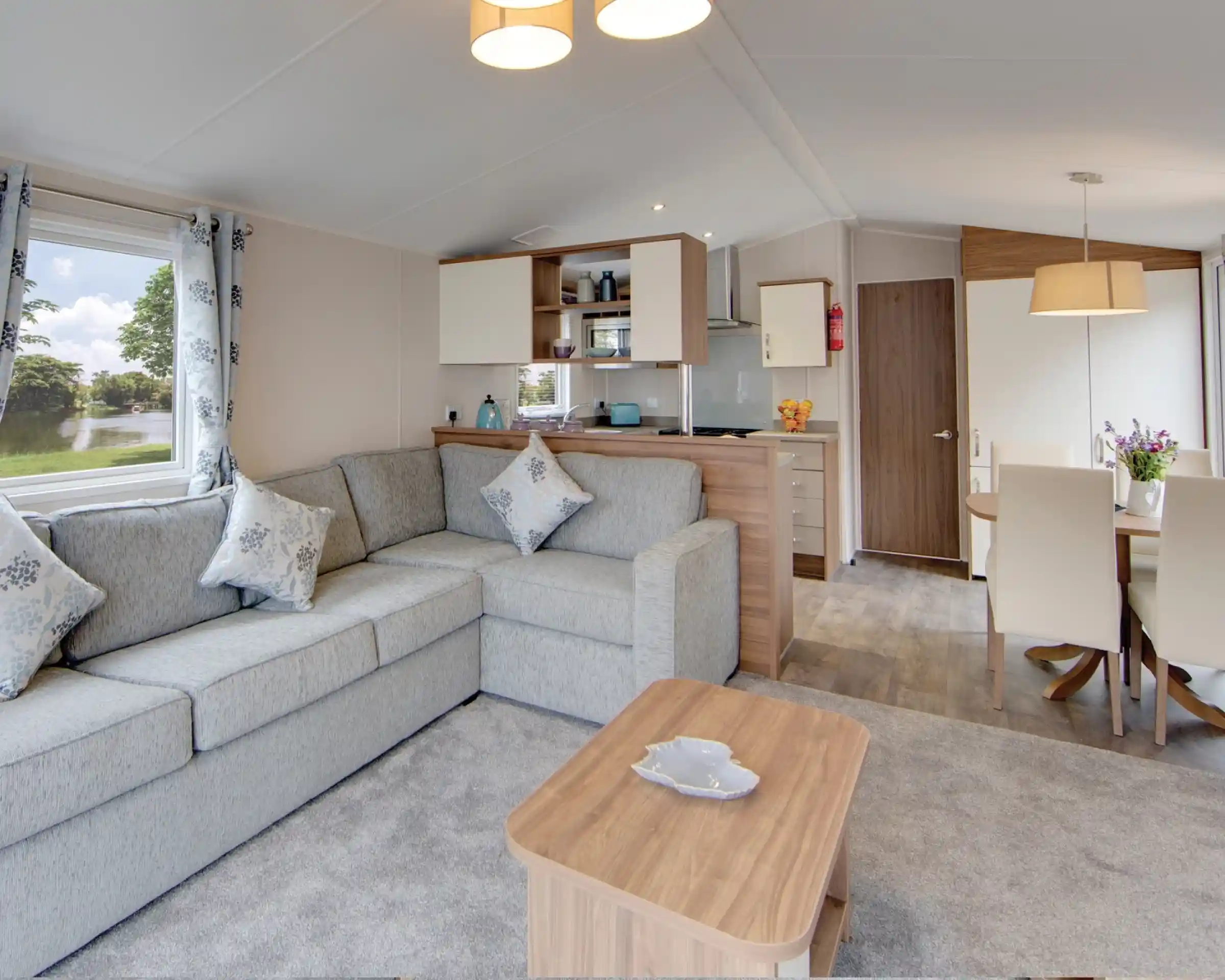 Interior view of a modern living area with an L-shaped grey sofa, coffee table, kitchen counter, and dining table with chairs.