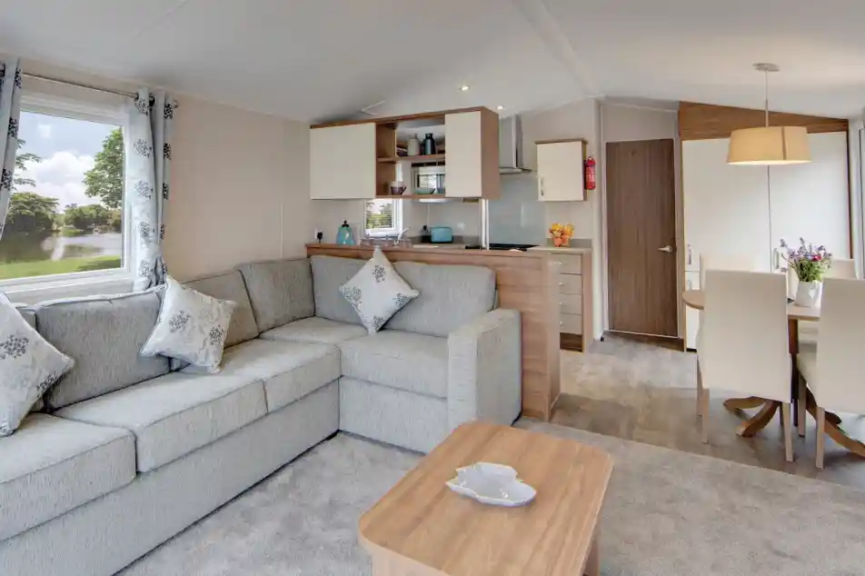 Interior view of a modern living area with an L-shaped grey sofa, coffee table, kitchen counter, and dining table with chairs.