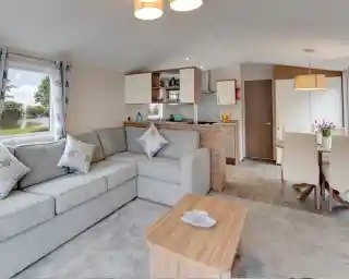 Interior view of a modern living area with an L-shaped grey sofa, coffee table, kitchen counter, and dining table with chairs.