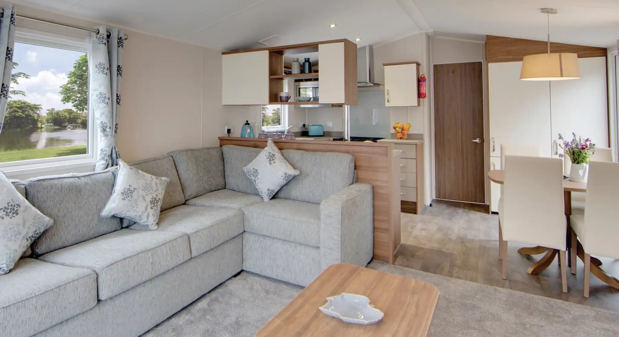 Interior view of a modern living area with an L-shaped grey sofa, coffee table, kitchen counter, and dining table with chairs.
