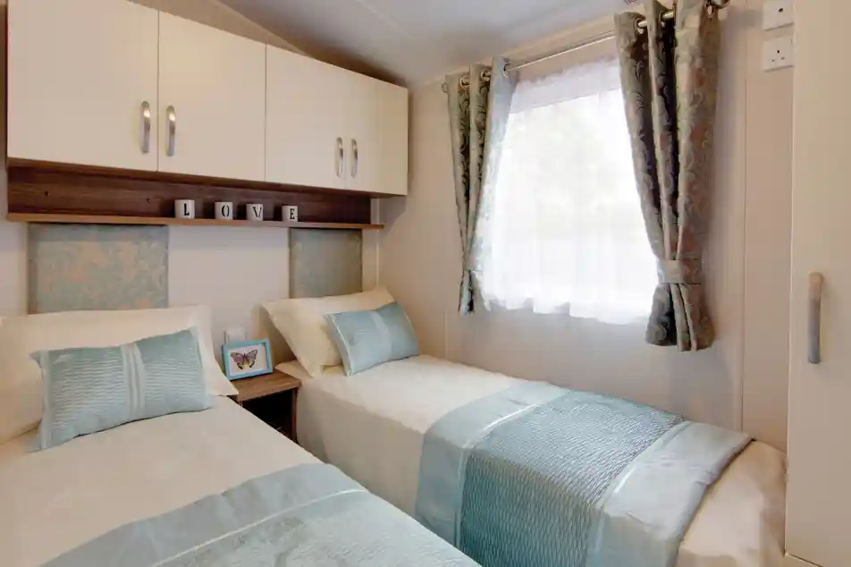 A cosy twin bedroom with two single beds dressed in cream bedding and light blue throws and cushions. Each bed has a patterned blue headboard, and a small wooden nightstand with a butterfly picture frame sits between them. Above the beds are white cabinets and a shelf with decorative blocks spelling “LOVE.” A window with sheer white curtains and patterned drapes lets in natural light.