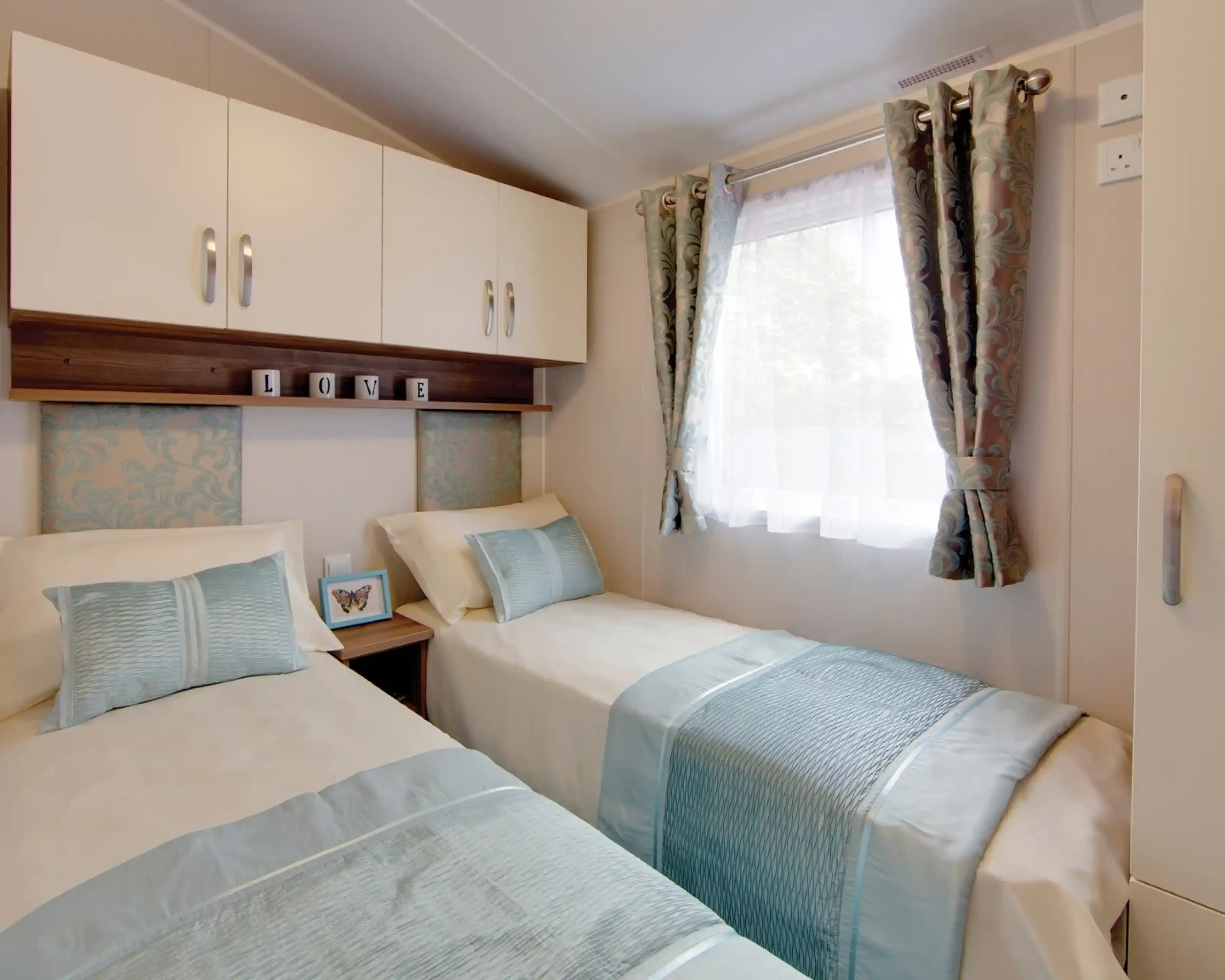 A cosy twin bedroom with two single beds dressed in cream bedding and light blue throws and cushions. Each bed has a patterned blue headboard, and a small wooden nightstand with a butterfly picture frame sits between them. Above the beds are white cabinets and a shelf with decorative blocks spelling “LOVE.” A window with sheer white curtains and patterned drapes lets in natural light.