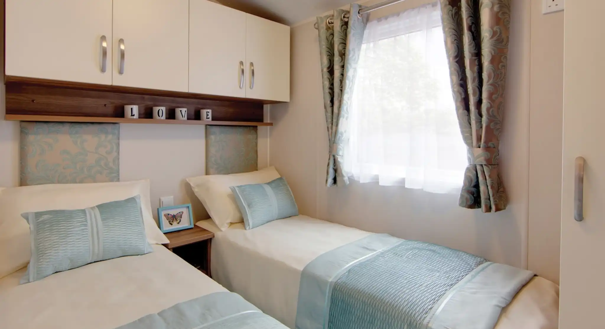 A cosy twin bedroom with two single beds dressed in cream bedding and light blue throws and cushions. Each bed has a patterned blue headboard, and a small wooden nightstand with a butterfly picture frame sits between them. Above the beds are white cabinets and a shelf with decorative blocks spelling “LOVE.” A window with sheer white curtains and patterned drapes lets in natural light.