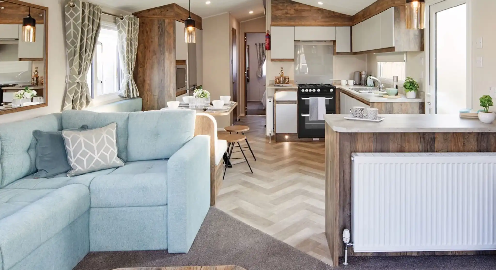 A spacious living room featuring a couch and kitchen area, furnished with a coffee table and pillows. The interior showcases hardwood flooring and laminate accents, with cabinetry and home appliances in the kitchen section. Bright windows allow natural light to illuminate the space, creating a light atmosphere.