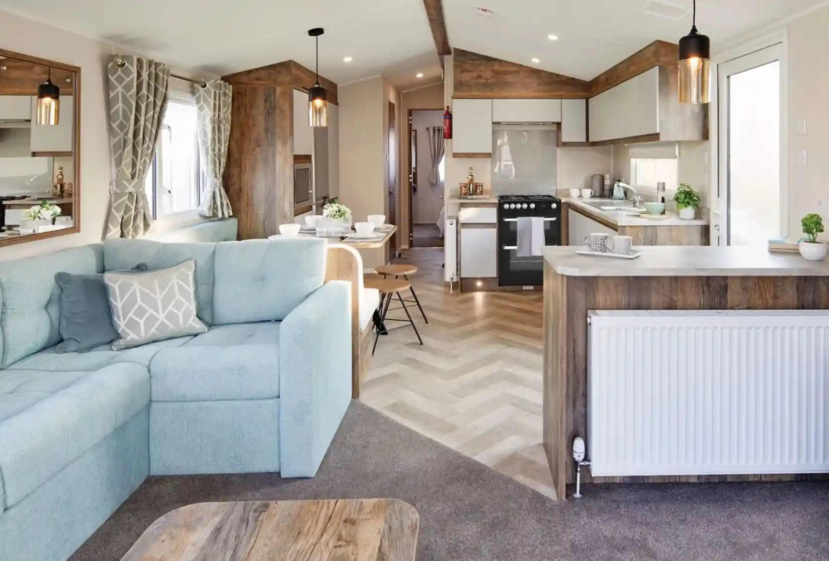 A spacious living room featuring a couch and kitchen area, furnished with a coffee table and pillows. The interior showcases hardwood flooring and laminate accents, with cabinetry and home appliances in the kitchen section. Bright windows allow natural light to illuminate the space, creating a light atmosphere.