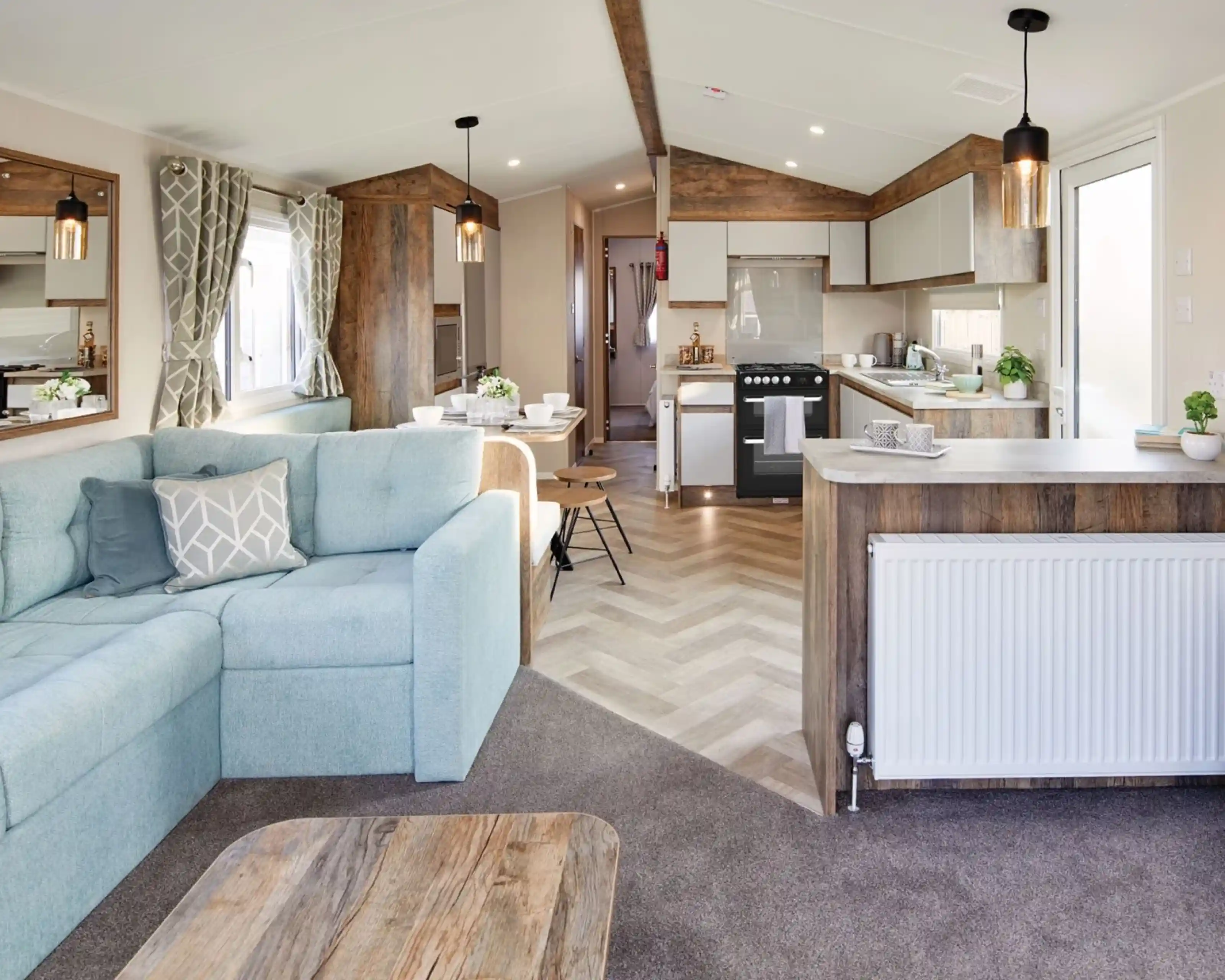 A spacious living room featuring a couch and kitchen area, furnished with a coffee table and pillows. The interior showcases hardwood flooring and laminate accents, with cabinetry and home appliances in the kitchen section. Bright windows allow natural light to illuminate the space, creating a light atmosphere.