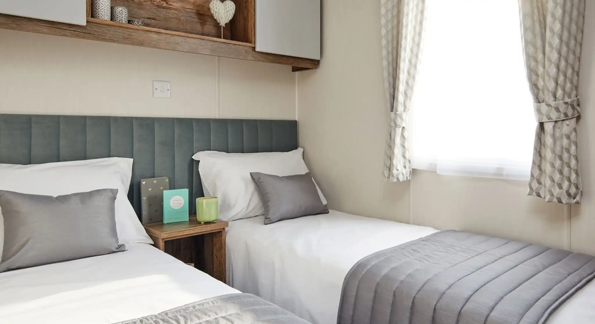 A bedroom with twin beds, a plush headboard, and bedside table, together with overhead storage and a window with curtains.