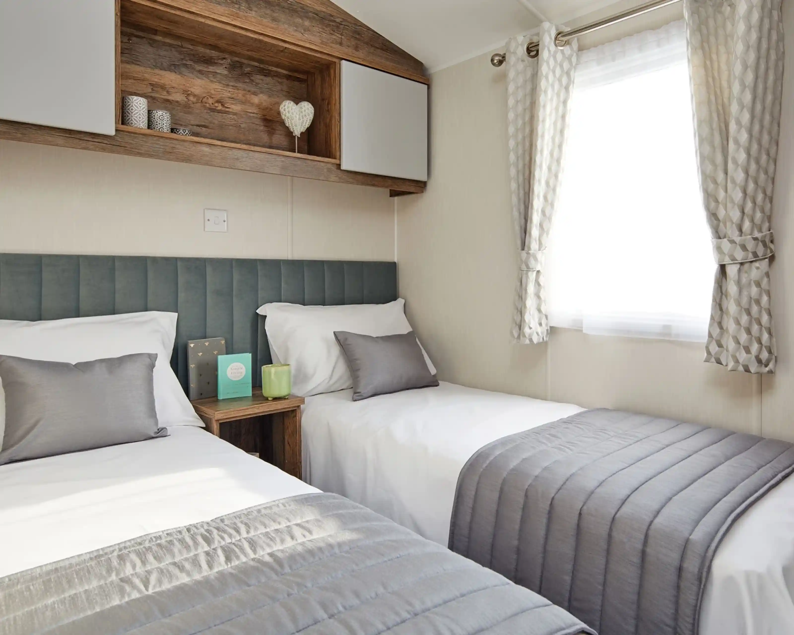 A bedroom with twin beds, a plush headboard, and bedside table, together with overhead storage and a window with curtains.