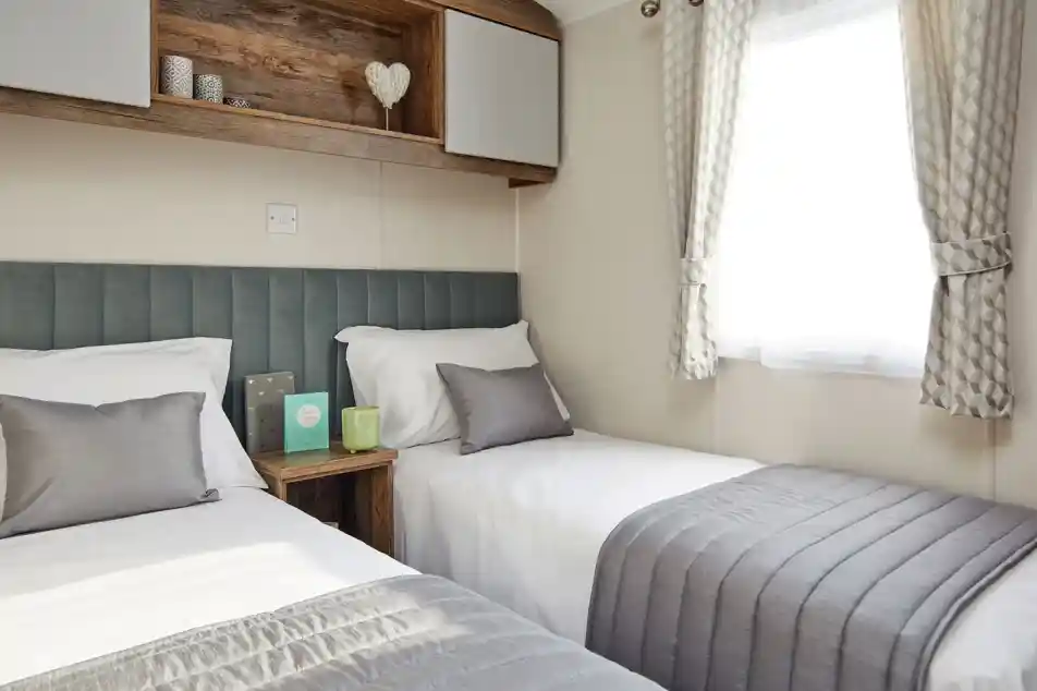 A bedroom with twin beds, a plush headboard, and bedside table, together with overhead storage and a window with curtains.