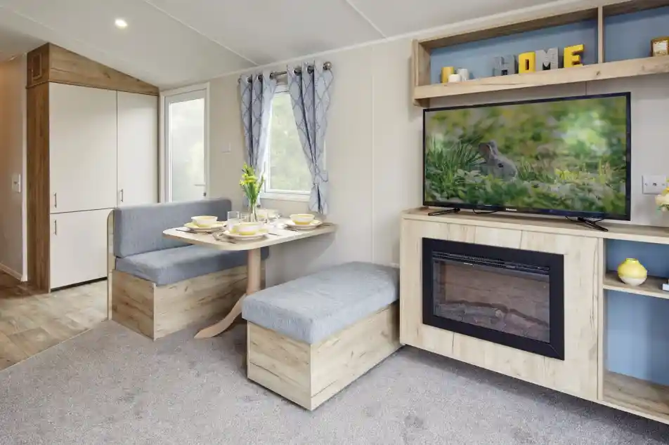 A cozy interior of a modern living space featuring a table set for a meal, gray benches, a mounted TV displaying a rabbit, and a decorative fireplace. Light streams in through large windows, highlighting the warm wooden accents and soft carpet.