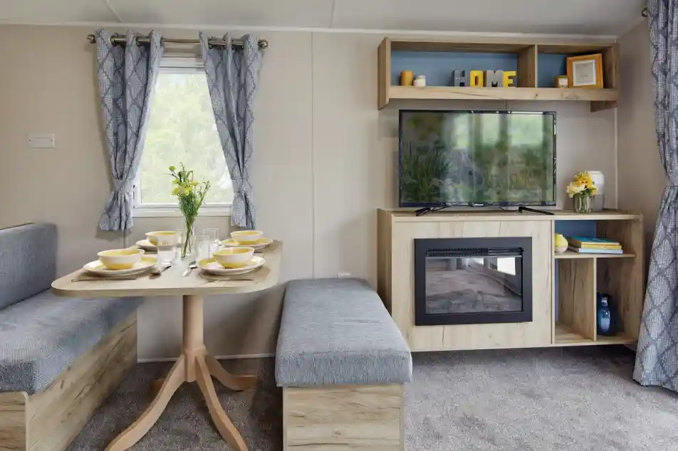 A cozy living space featuring a wooden dining table set for four, with grey benches and a decorative vase of flowers. The area has a modern TV mounted on a wooden shelf, with shelves displaying various decor items, and light curtains framing two windows. A faux fireplace adds warmth to the ambiance.