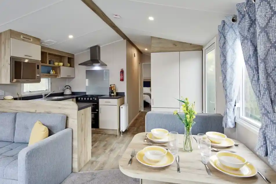 A cozy interior of a modern mobile home featuring a living area with a gray sofa, a dining table set with yellow dishes, and a kitchenette with a stove and refrigerator. Light floods in through large windows, and a bouquet of flowers adds a touch of color to the scene.