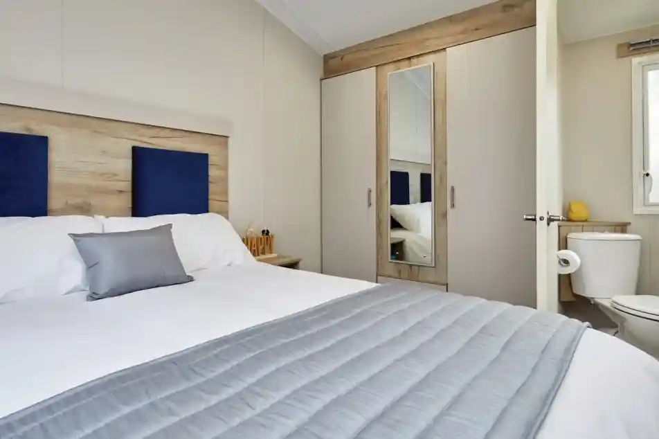 A cozy bedroom featuring a queen-sized bed with a gray blanket and decorative pillows. A wardrobe with a mirror is visible, along with a small bathroom area. Neutral-colored walls create a warm atmosphere.