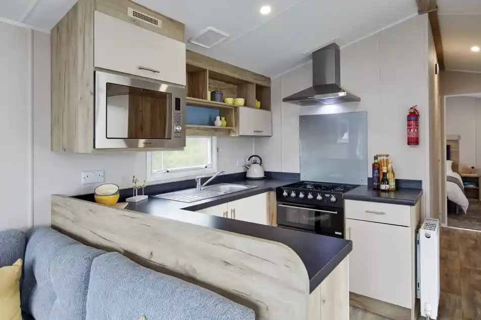 Modern kitchen with a blue sofa, featuring wooden cabinets, a stainless steel microwave, a blue glass backsplash, and a gas stove with an oven. Blue and yellow decor elements add a cozy touch.