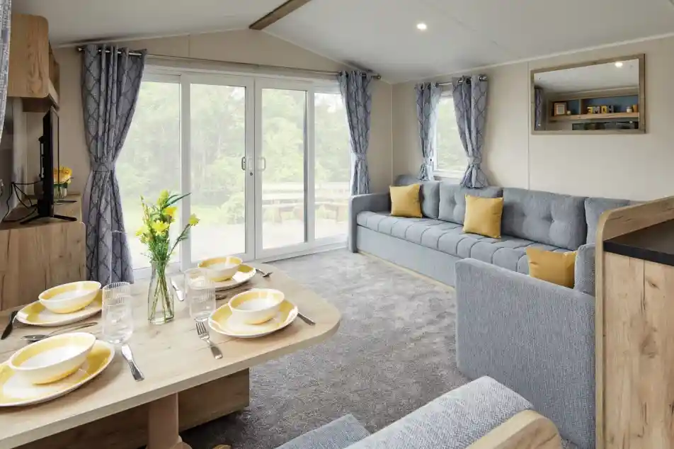 A modern living space featuring a gray sofa adorned with yellow cushions, a light wooden dining table set for four with yellow plates, and large glass doors that open to a patio area. Curtains with a blue pattern frame the windows, and there are fresh flowers on the table.