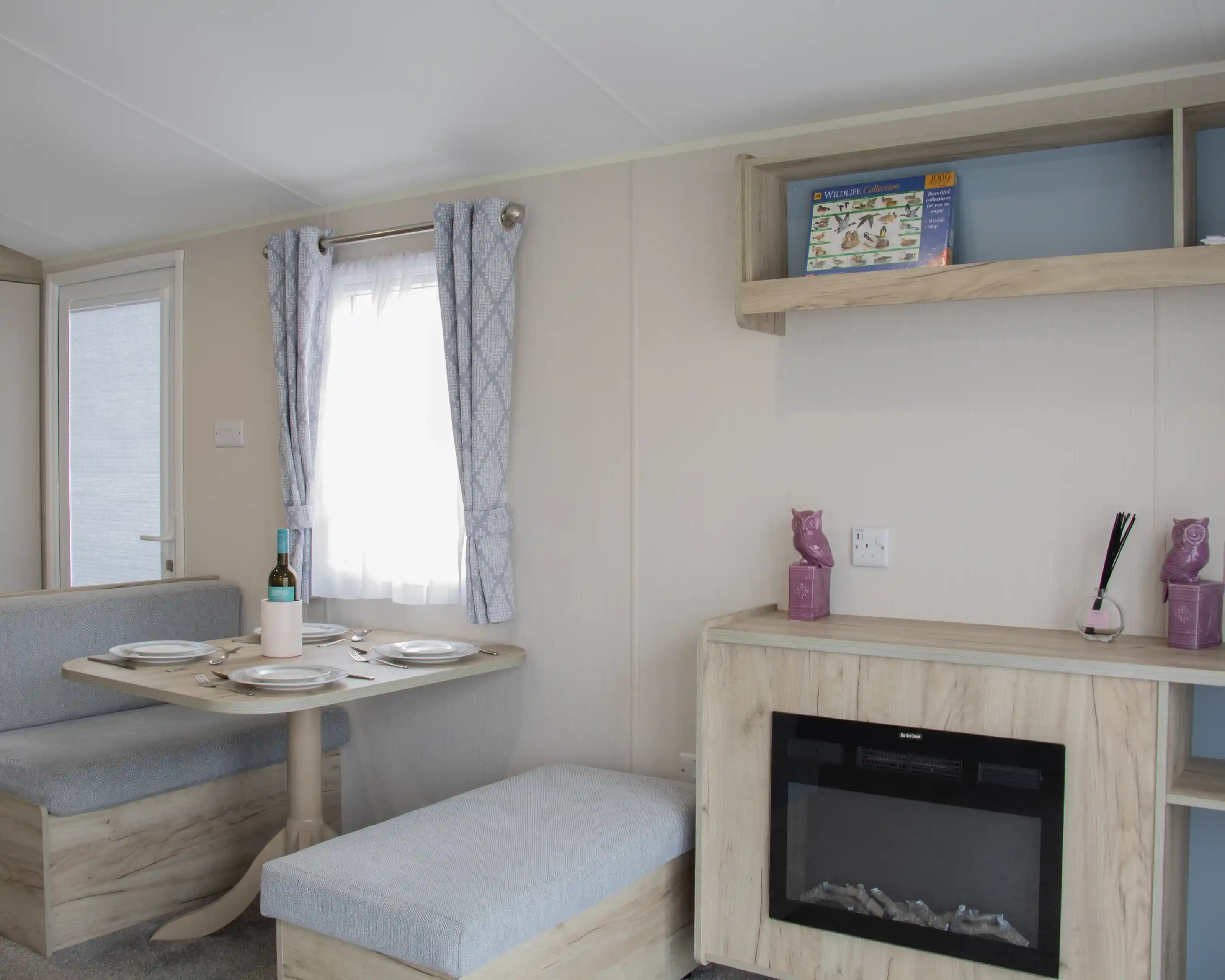 A cozy interior of a modern caravan featuring a dining area with a round table set for two, a small sofa, and a decorative shelf. A faux fireplace adds warmth to the space, and light curtains allow natural light to enter through a window.