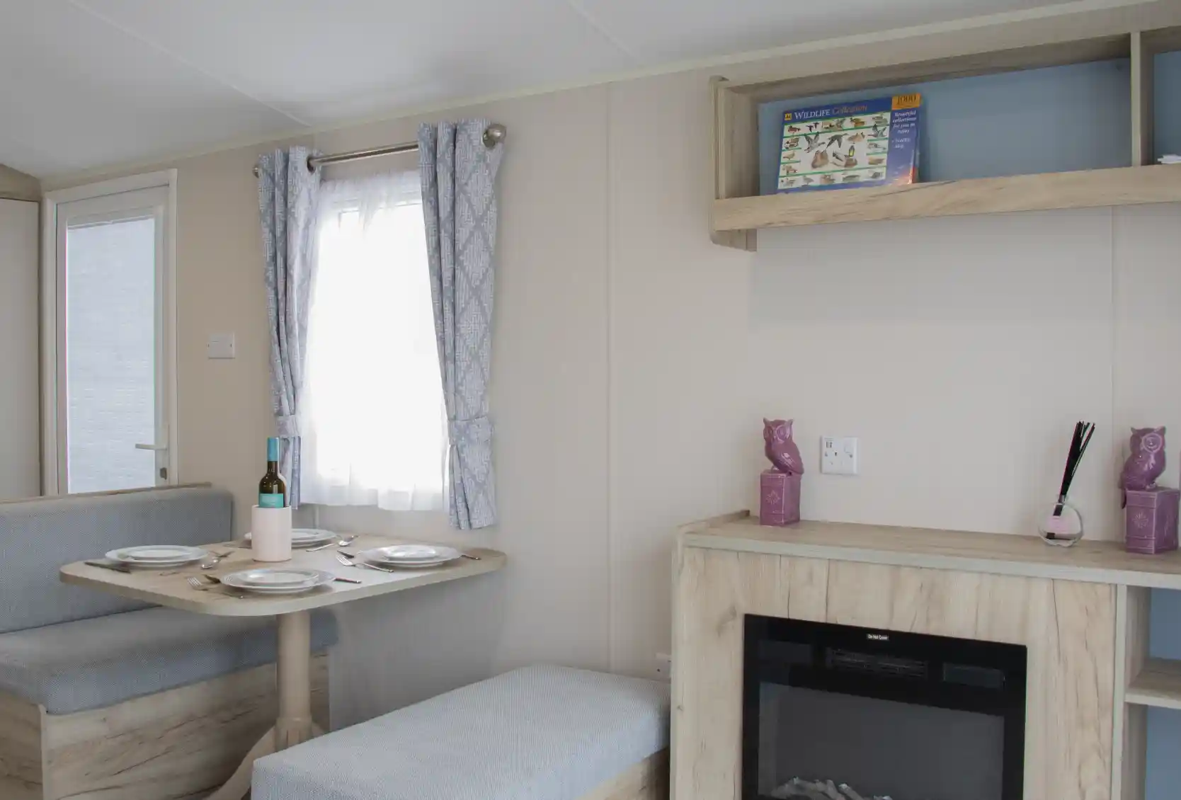 A cozy interior of a modern caravan featuring a dining area with a round table set for two, a small sofa, and a decorative shelf. A faux fireplace adds warmth to the space, and light curtains allow natural light to enter through a window.