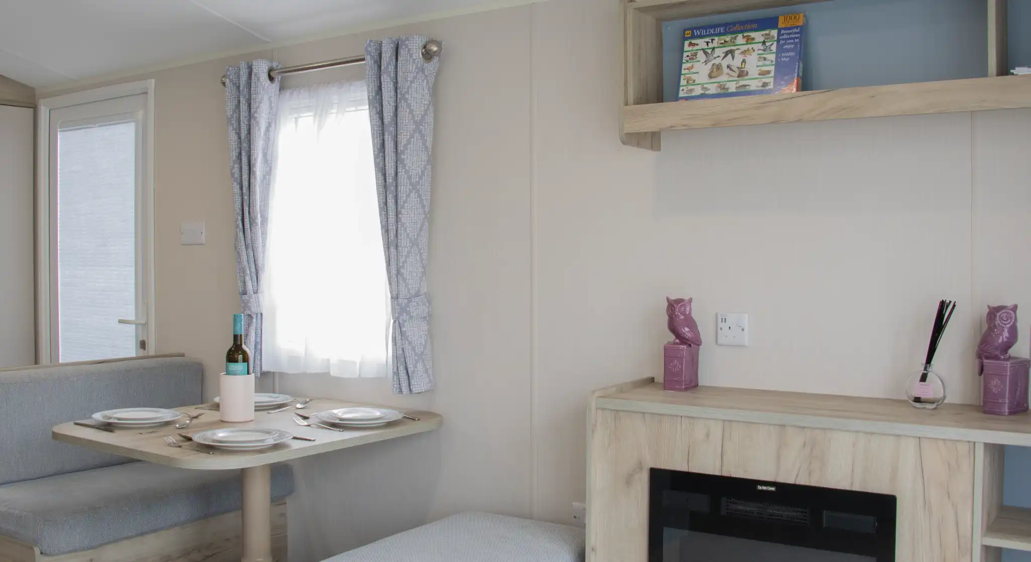A cozy interior of a modern caravan featuring a dining area with a round table set for two, a small sofa, and a decorative shelf. A faux fireplace adds warmth to the space, and light curtains allow natural light to enter through a window.
