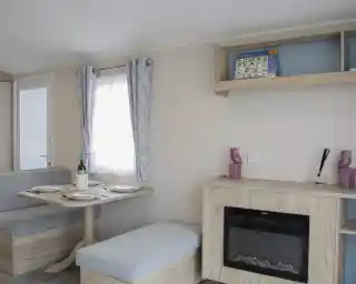 A cozy interior of a modern caravan featuring a dining area with a round table set for two, a small sofa, and a decorative shelf. A faux fireplace adds warmth to the space, and light curtains allow natural light to enter through a window.
