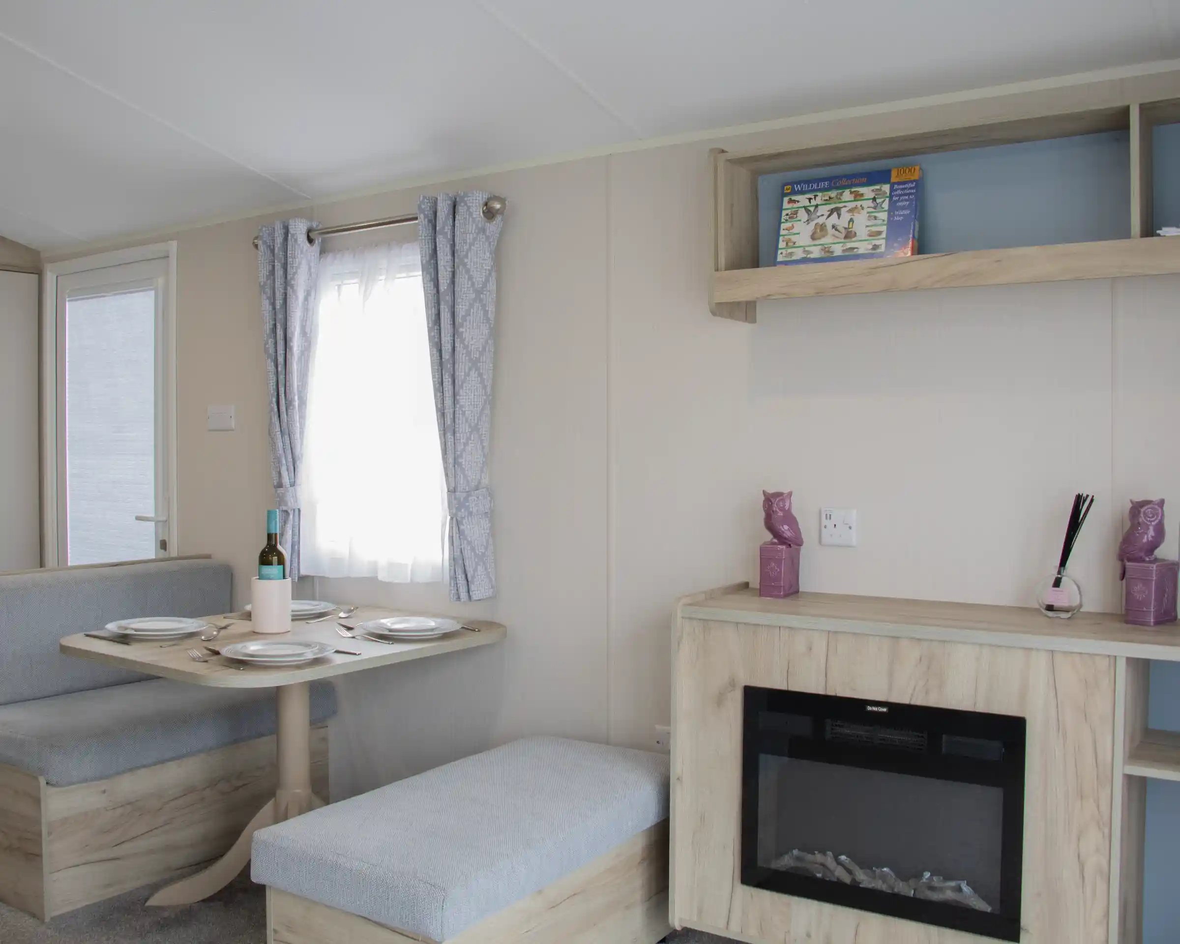 A cozy interior of a modern caravan featuring a dining area with a round table set for two, a small sofa, and a decorative shelf. A faux fireplace adds warmth to the space, and light curtains allow natural light to enter through a window.