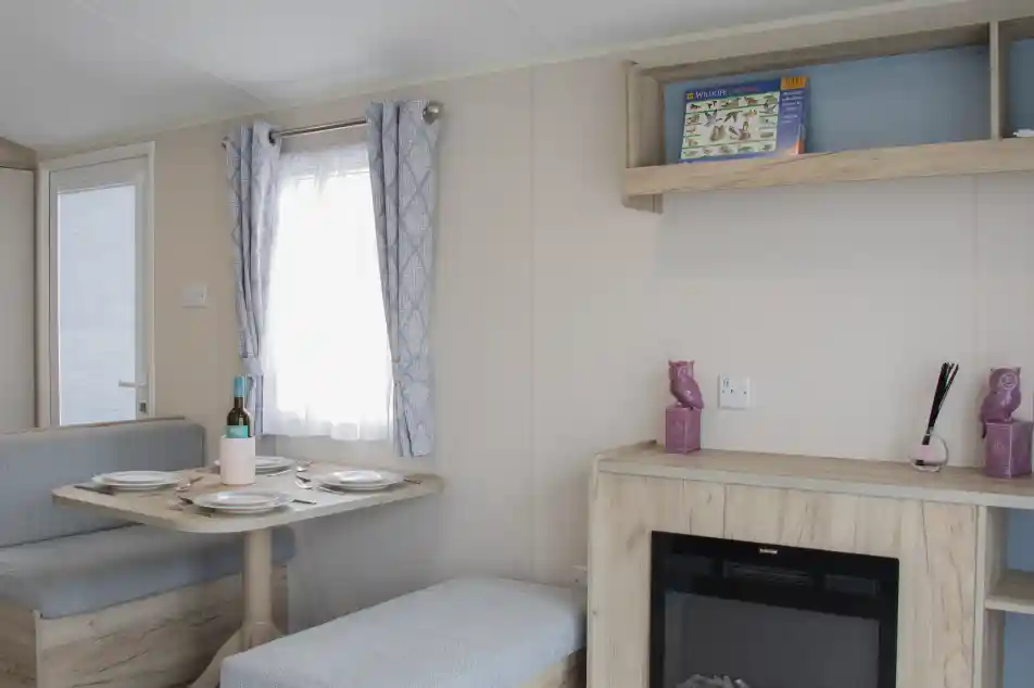 A cozy interior of a modern caravan featuring a dining area with a round table set for two, a small sofa, and a decorative shelf. A faux fireplace adds warmth to the space, and light curtains allow natural light to enter through a window.