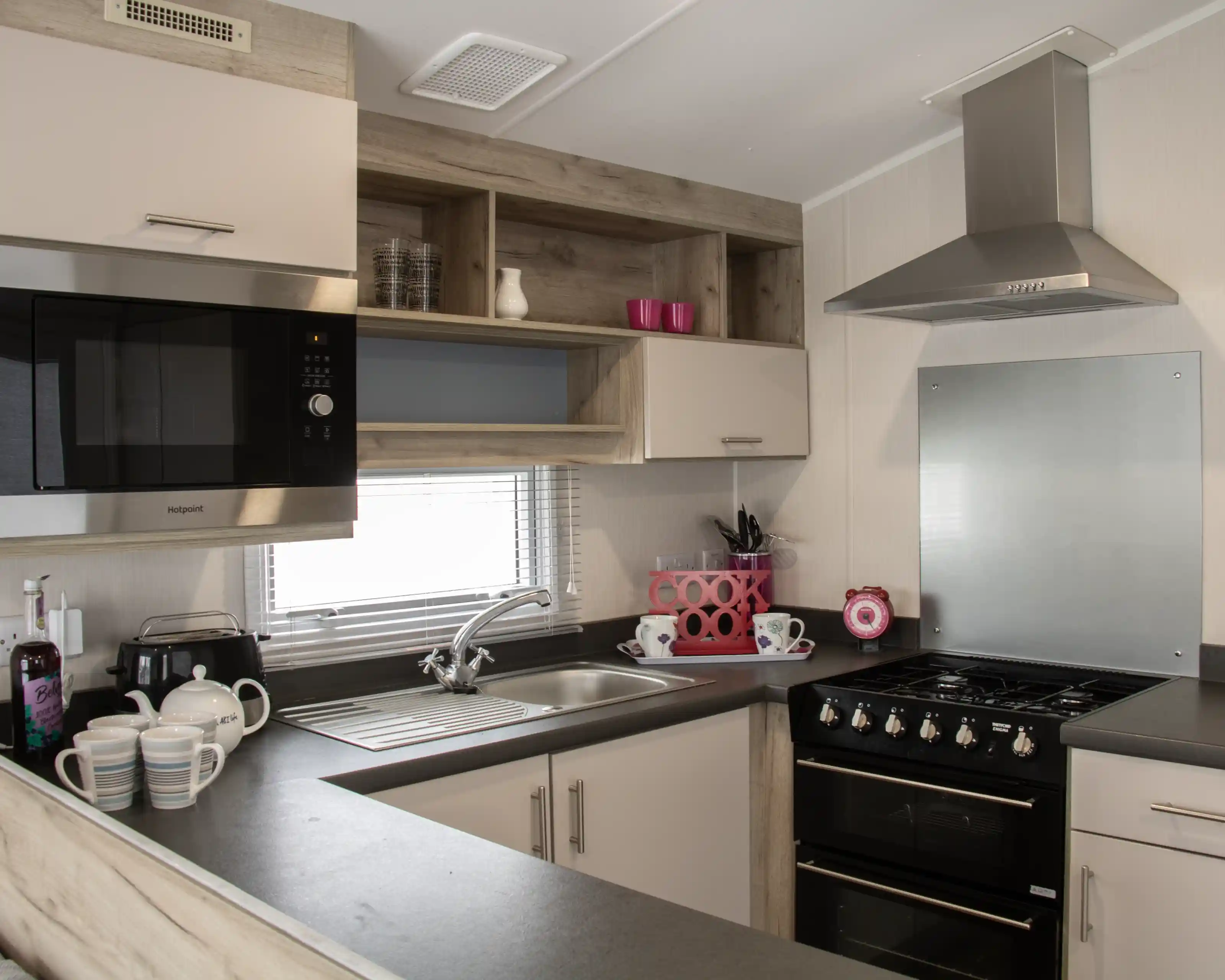 A modern kitchen featuring a silver microwave, a stainless steel sink, and a black gas stove. There are wooden shelves above with pink cups and kitchenware. The countertop is dark with white dishes and a teapot arranged neatly. A window provides natural light.