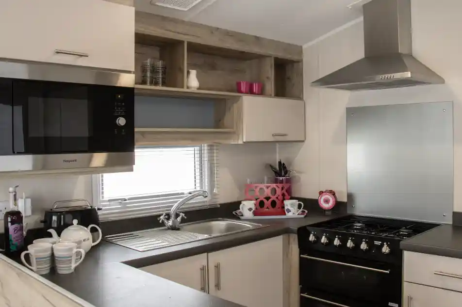 A modern kitchen featuring a silver microwave, a stainless steel sink, and a black gas stove. There are wooden shelves above with pink cups and kitchenware. The countertop is dark with white dishes and a teapot arranged neatly. A window provides natural light.