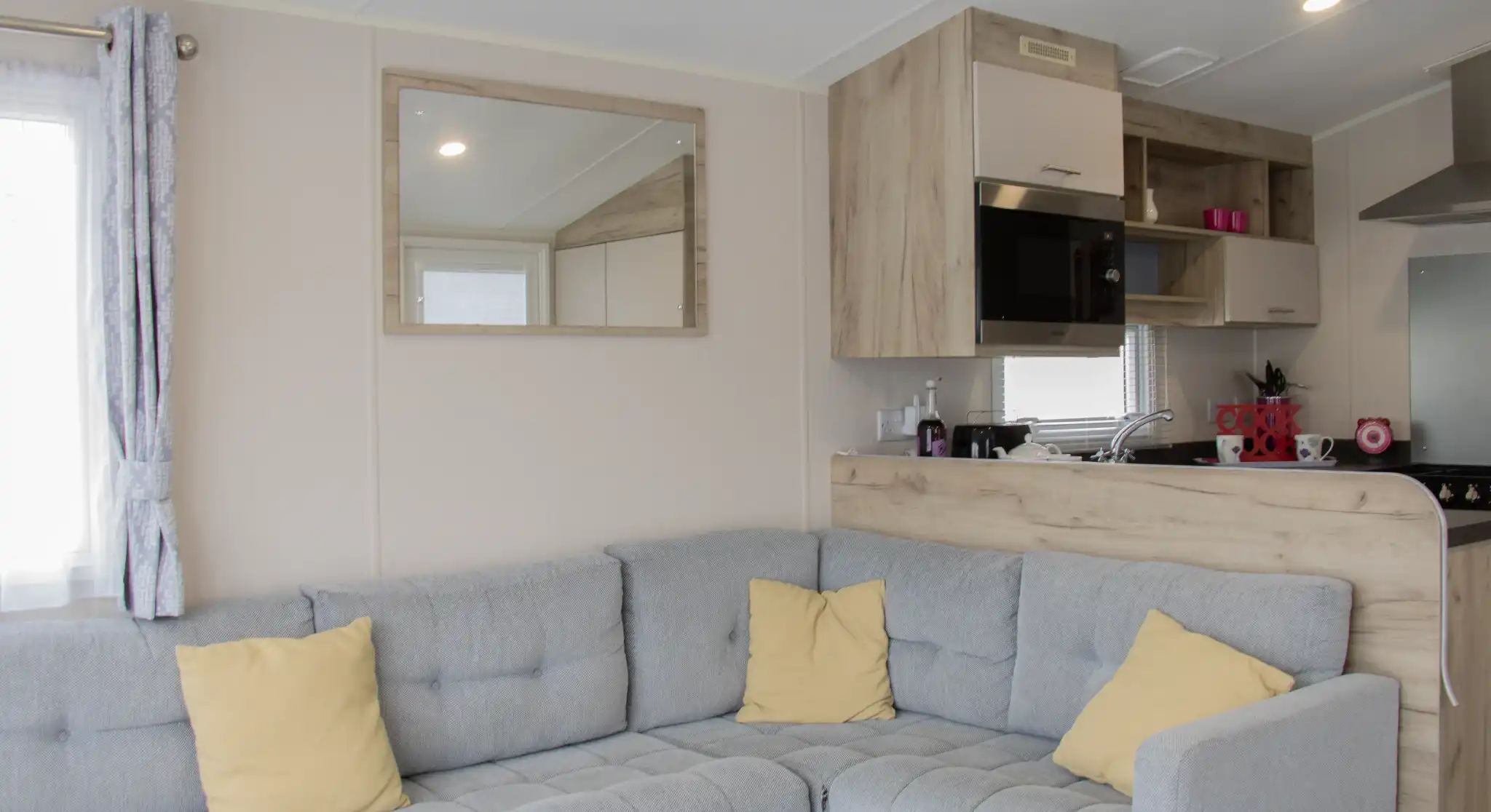 A cozy, modern interior of a mobile home featuring a gray sectional sofa with yellow cushions, a wooden kitchenette with a mirror, and a bright window.